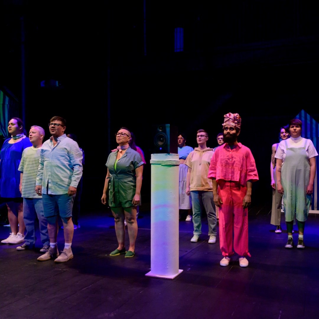 Photo of 12 choir singers on a black stage singer. The singers are spaced out on the stage. At the center of the stage is a round podium with a speaker on top. 