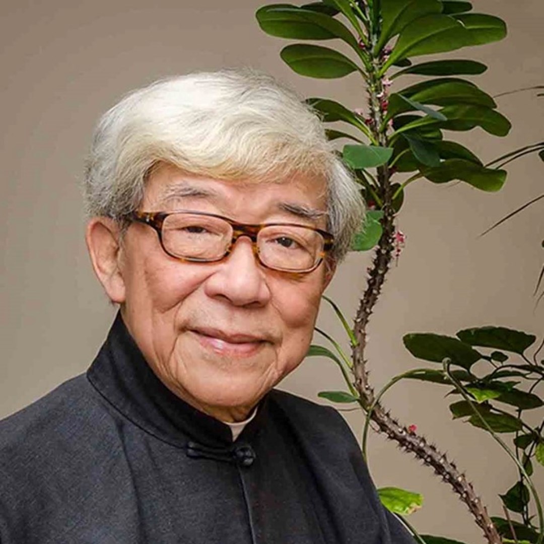 Man with white hair, glasses, and black traditional Chinese shirt. Tall green plant in background