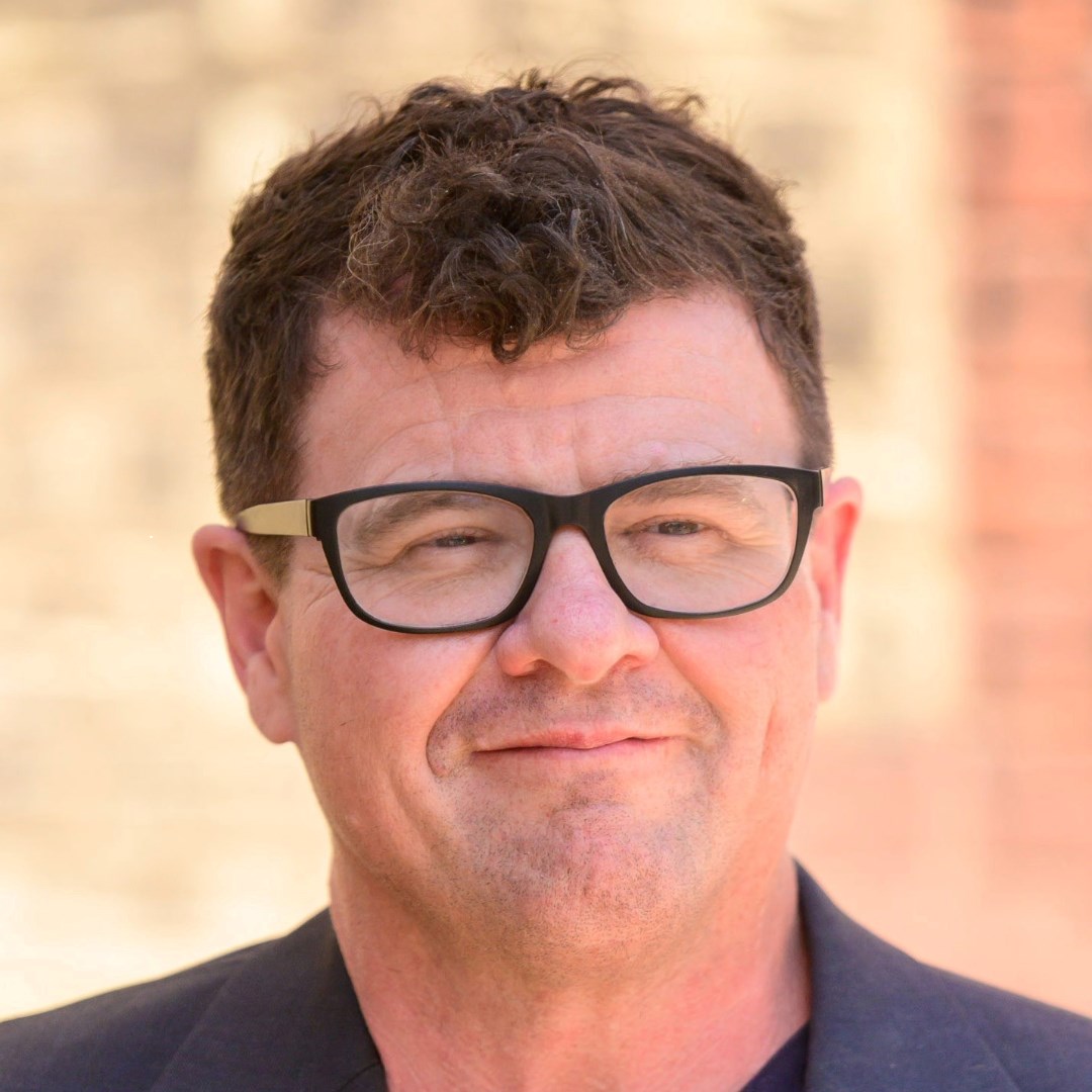 Headshot of transgender man with white skin, brown hair, black glasses, and black suit. Background is blurred and the colour beige. 